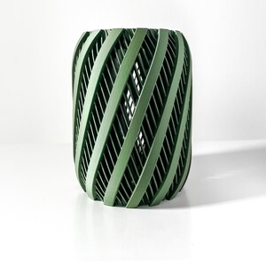 May include: A green, cylindrical planter with a unique, woven design. The planter is made of a durable material and has a modern, minimalist aesthetic.