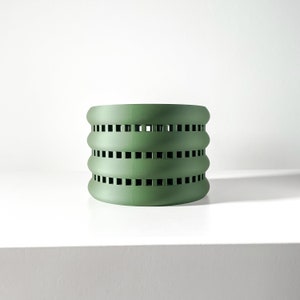 May include: A green cylindrical planter with a decorative pattern of square cutouts. The planter has four rows of cutouts.