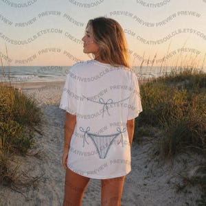 Pode incluir: Uma t-shirt branca de manga curta com um gr&aacute;fico de linhas azuis nas costas. O design mostra um biqu&iacute;ni de duas pe&ccedil;as com um top de atar e uma cueca, ambos representados num estilo de contorno azul simples e minimalista.