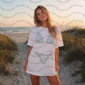 Pode incluir: Uma t-shirt branca de manga curta com um gr&aacute;fico de linhas azul-escuro de um biqu&iacute;ni de duas pe&ccedil;as na parte da frente. O gr&aacute;fico mostra um top de biqu&iacute;ni triangular com bordas franzidas e uma cueca a condizer com la&ccedil;os laterais.