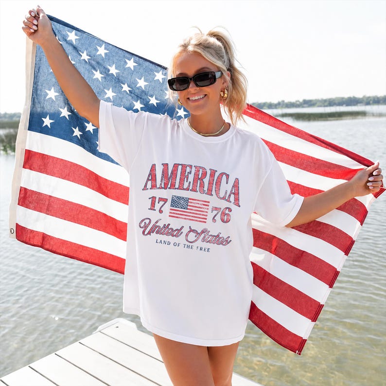 May include: A white short-sleeve t-shirt featuring a red and blue graphic print that reads AMERICA 1776 United States LAND OF THE FREE with a small American flag icon. The shirt is worn in front of a large, horizontal American flag held up by arms.
