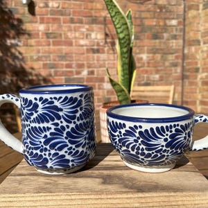 May include: Two ceramic mugs with a blue and white floral pattern. The larger mug has a handle and is taller than the smaller mug. The smaller mug has a handle and is wider than it is tall. Both mugs are on a wooden surface.