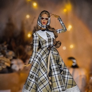 May include: A fashion doll wearing a white collared dress with a plaid pattern in shades of brown, black, and white. The doll is wearing white sunglasses and has brown hair. The dress has a lace trim and the doll is wearing white heels.