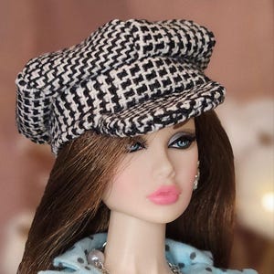Puede incluir: Una muñeca con una gorra de estilo newsboy a cuadros blancos y negros. La muñeca tiene cabello castaño largo, ojos azules y lápiz labial rosa. Lleva una blusa celeste con lunares y un collar de perlas.