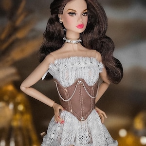May include: A fashion doll with brown hair and a white lace dress with a brown corset. The doll is wearing a choker necklace and earrings. The dress has gold bows and the doll has pink lipstick.