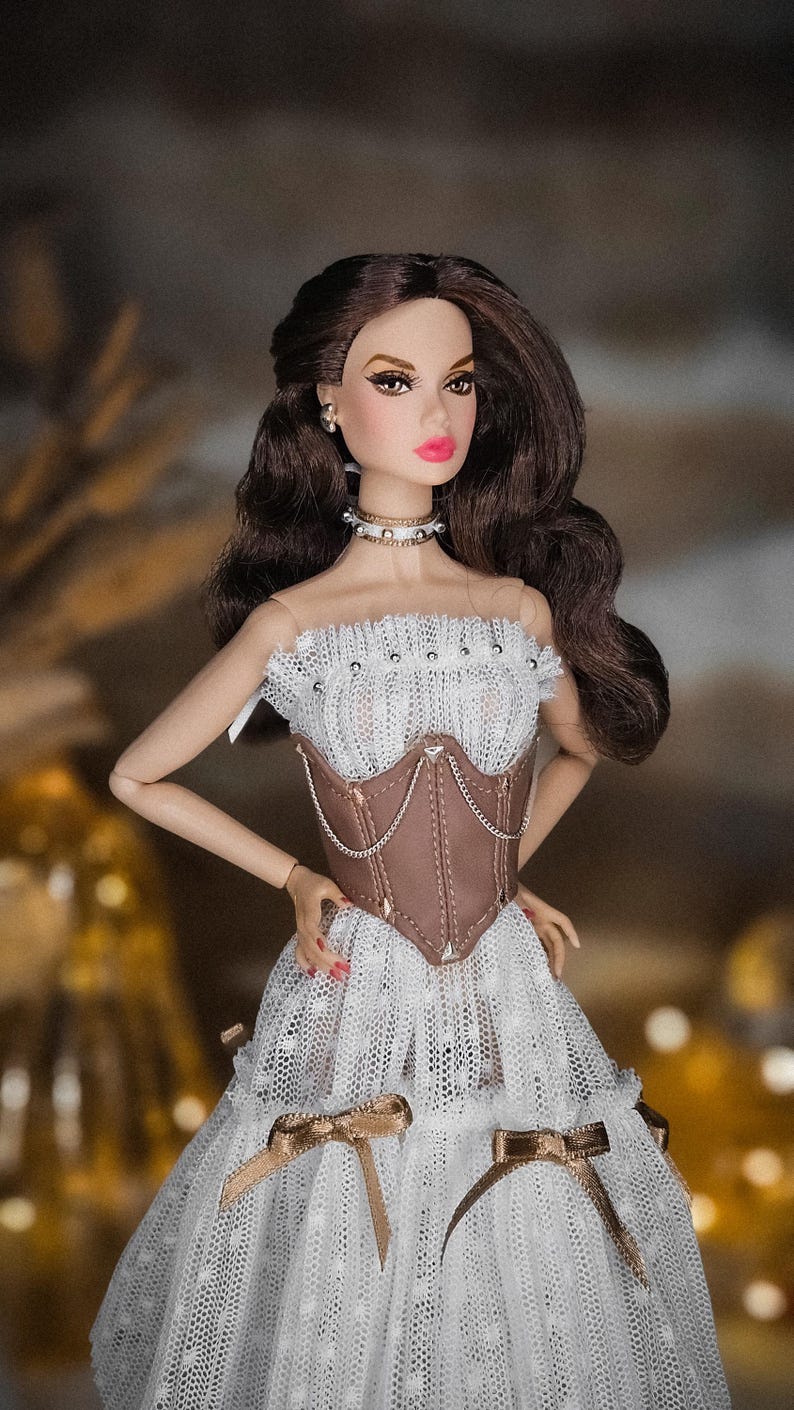 May include: A fashion doll with brown hair and a white lace dress with a brown corset. The doll is wearing a choker necklace and earrings. The dress has gold bows and the doll has pink lipstick.