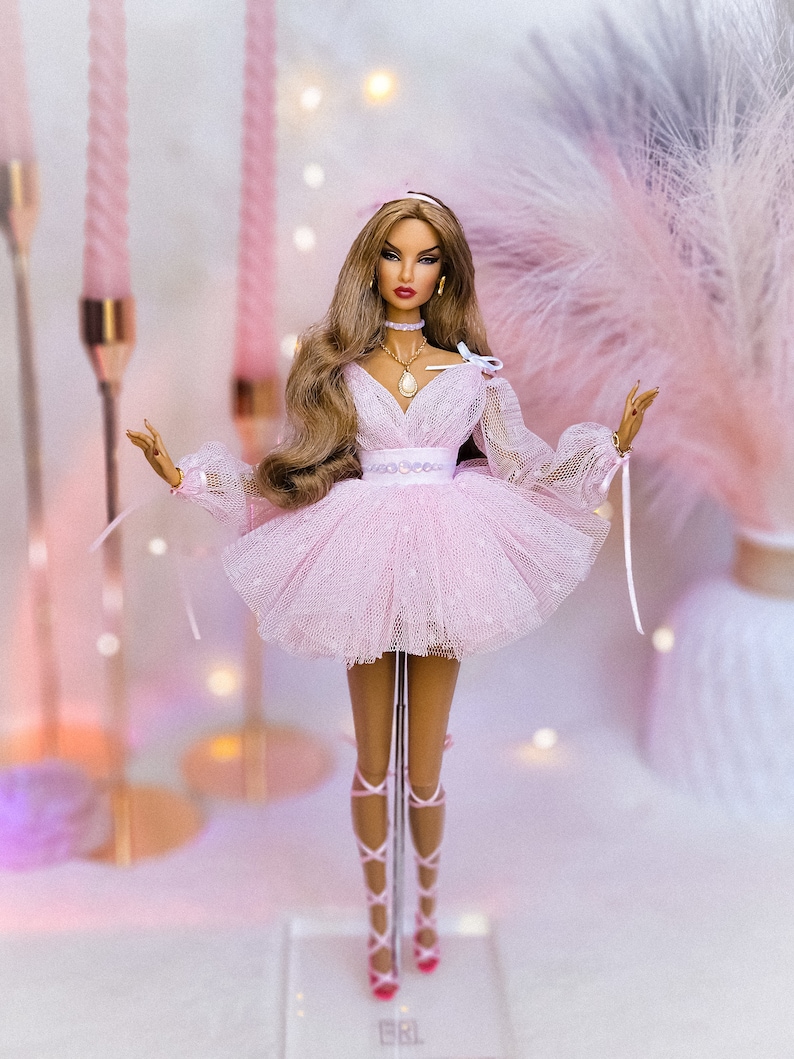 May include: A pink tulle dress with a white lace overlay and a pearl embellishment. The doll is wearing a gold necklace with a pearl pendant and a pink headband. The doll is standing on a clear display stand.