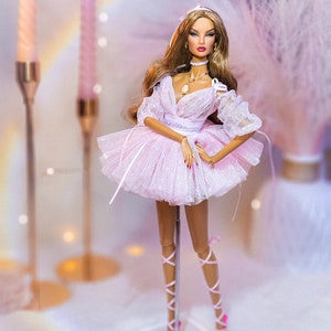 May include: A fashion doll with long brown hair wearing a pink tulle dress and pink shoes. The doll is standing on a clear plastic stand.