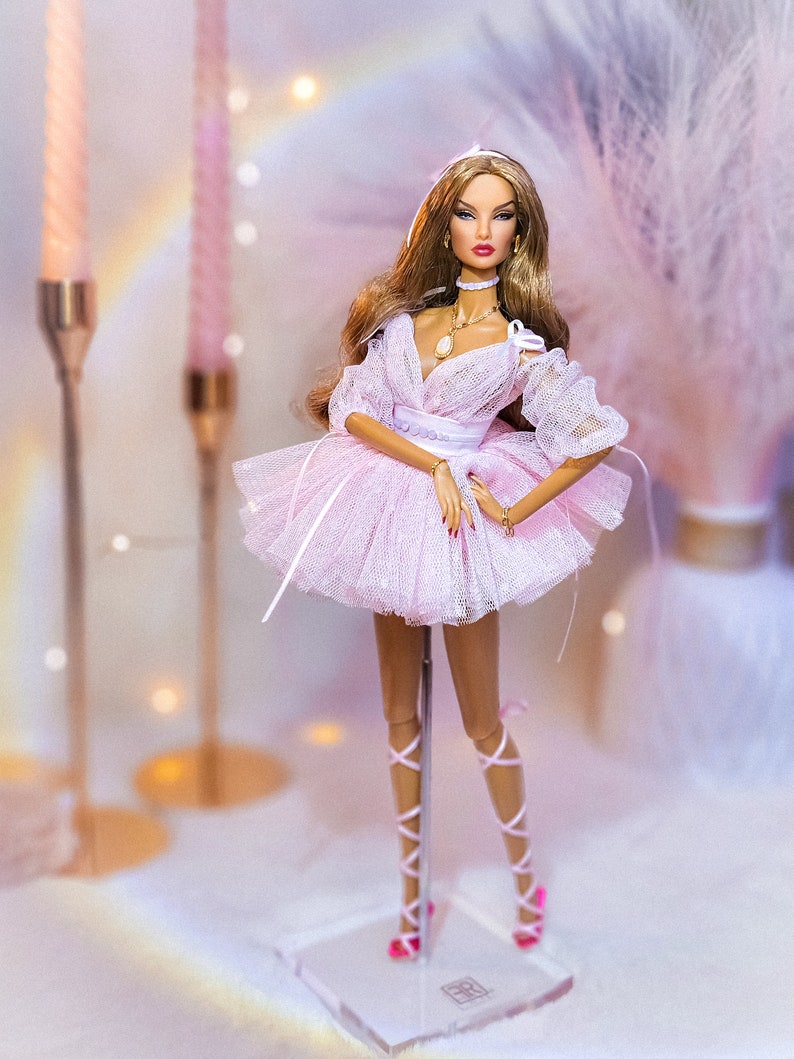 May include: A fashion doll with long brown hair wearing a pink tulle dress and pink shoes. The doll is standing on a clear plastic stand.