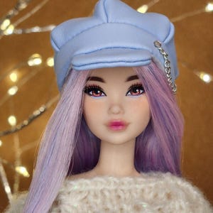Könnte beinhalten: Eine Puppe mit lavendelfarbenem Haar und einer hellblauen Mütze mit silberner Kettendetail. Die Puppe trägt einen weißen, strukturierten Pullover. Die Puppe hat rosa Lidschatten und rosa Lippenstift.