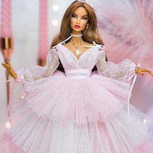 May include: A doll wearing a pink tulle dress with a white underskirt. The doll has long brown hair, red lips, and is wearing a gold necklace with a pendant.