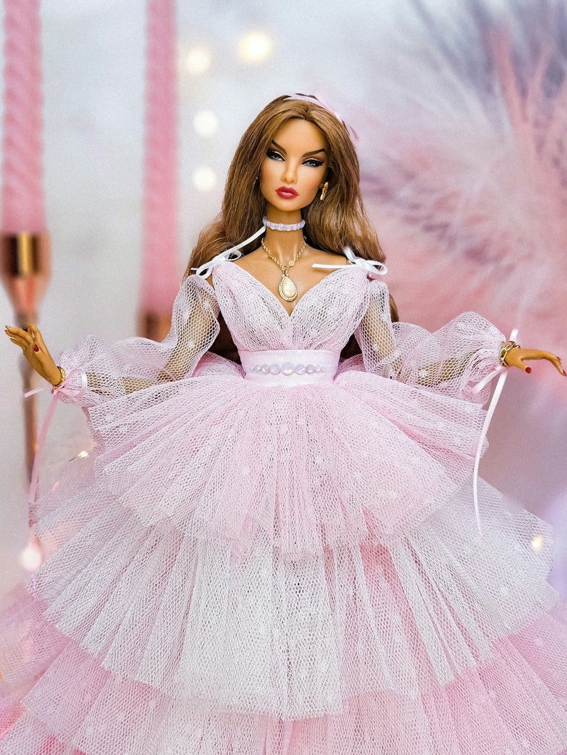 May include: A doll wearing a pink tulle dress with a white underskirt. The doll has long brown hair, red lips, and is wearing a gold necklace with a pendant.