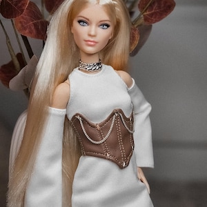 May include: A fashion doll with long blonde hair and blue eyes, wearing a white dress with long sleeves and a brown corset. The doll is accessorized with a silver chain necklace. The background includes a vase with brown leaves.