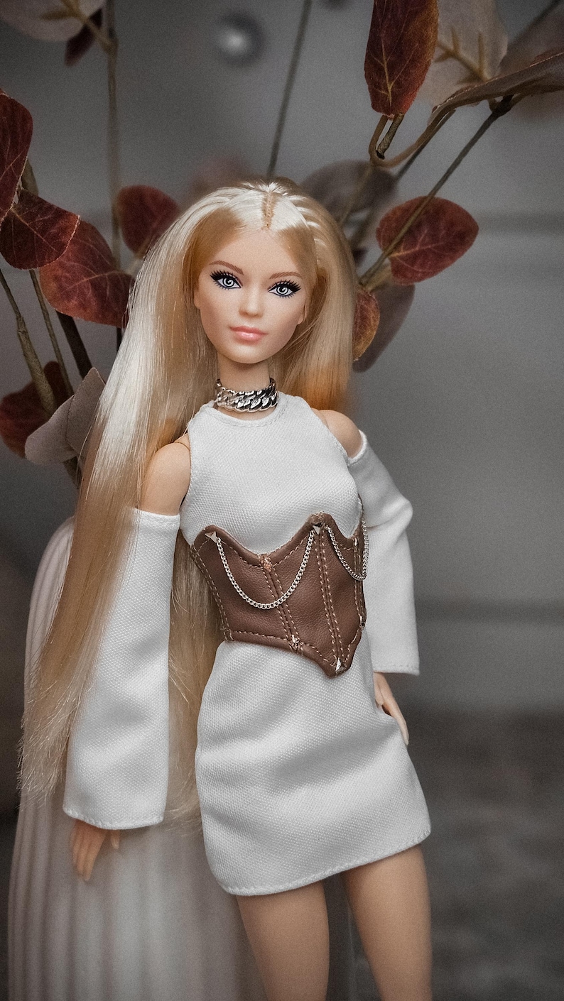 May include: A fashion doll with long blonde hair and blue eyes, wearing a white dress with long sleeves and a brown corset. The doll is accessorized with a silver chain necklace. The background includes a vase with brown leaves.
