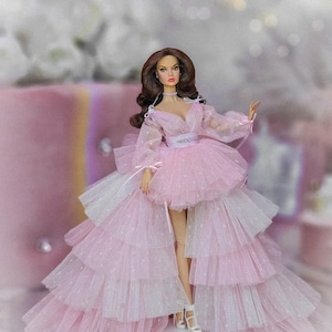 Könnte beinhalten: Ein rosa Tüllkleid mit einem weißen Tüllüberzug und einem taillierten Oberteil. Das Kleid hat einen mehrlagigen Rock und ist mit kleinen weißen Punkten verziert. Die Puppe trägt weiße Absätze.