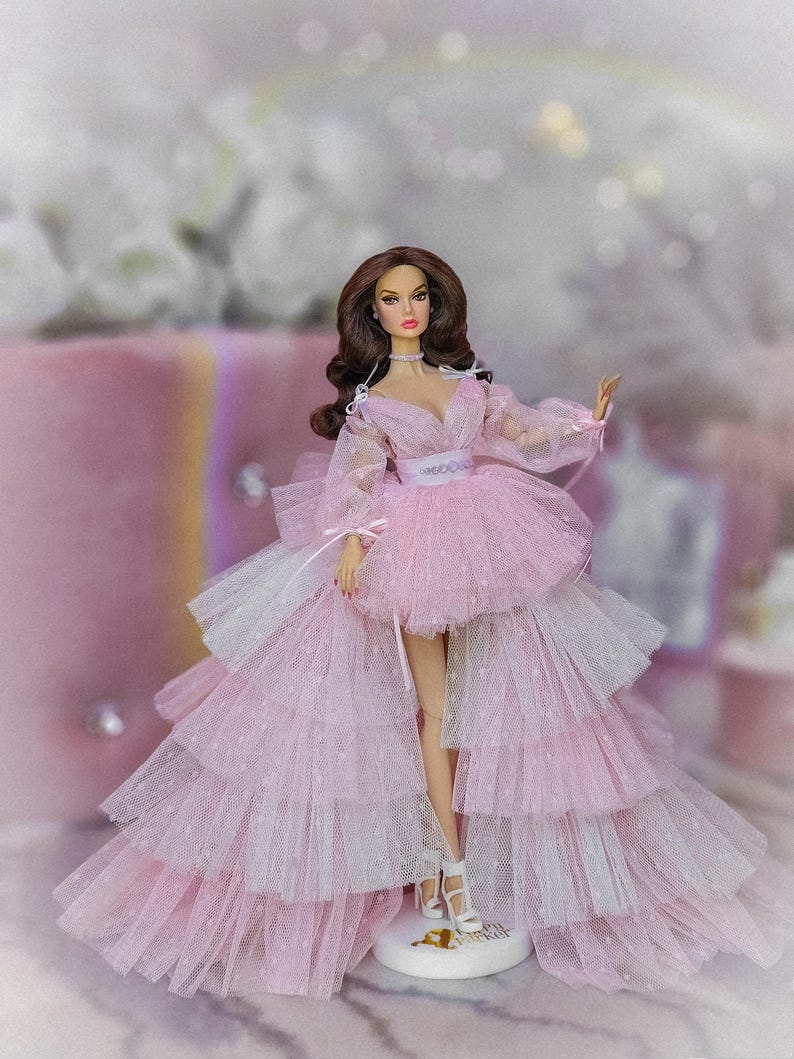 May include: A pink tulle dress with a white tulle overlay and a fitted bodice. The dress has a layered skirt and is decorated with small white dots. The doll is wearing white heels.