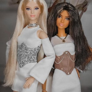 Puede incluir: Dos muñecas de moda posan juntas, cada una con un vestido blanco. Una muñeca tiene pelo largo rubio y un collar plateado, la otra tiene pelo oscuro y un corsé marrón. Los vestidos tienen detalles únicos en el corpiño.
