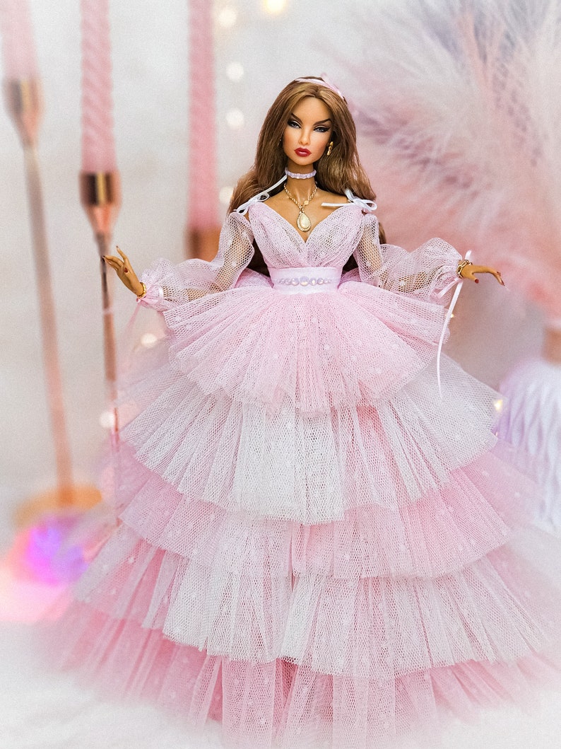 May include: A doll wearing a pink and white layered tulle dress with long sleeves and a sweetheart neckline. The doll has long brown hair, red lipstick, and is wearing a gold necklace with a pendant.