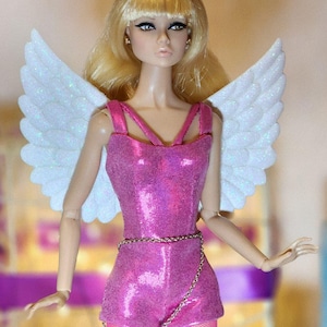 May include: A fashion doll with blonde hair and a pink sequined outfit, complete with white glittery wings. The doll is wearing a gold chain belt and pink fishnet stockings. The doll has a neutral expression.