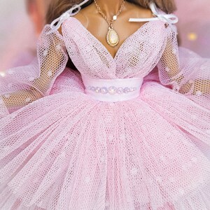 May include: A pink tulle dress with a sweetheart neckline and a white ribbon detail at the shoulders. The dress has a beaded belt and a delicate gold necklace with a pearlescent pendant.