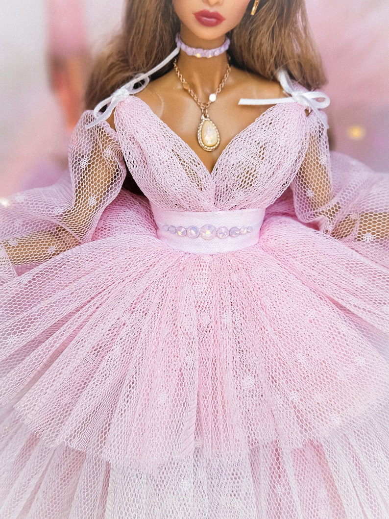 May include: A pink tulle dress with a sweetheart neckline and a white ribbon detail at the shoulders. The dress has a beaded belt and a delicate gold necklace with a pearlescent pendant.