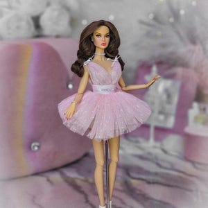 May include: A pink tulle dress with a sweetheart neckline and a white ribbon around the waist. The doll is wearing white heels and is standing on a white stand with the words "Dream Maker" printed on it.