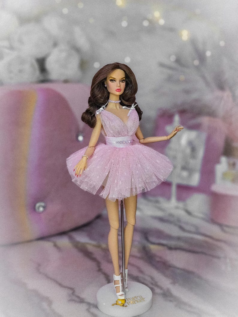 May include: A pink tulle dress with a sweetheart neckline and a white ribbon around the waist. The doll is wearing white heels and is standing on a white stand with the words "Dream Maker" printed on it.