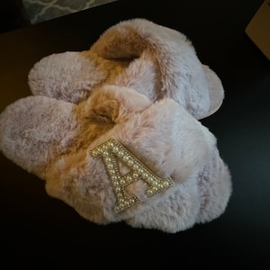 Peut inclure: Une paire de pantoufles moelleuses de couleur rose pâle avec une grande lettre "A" incrustée de perles sur le dessus d'une pantoufle.
