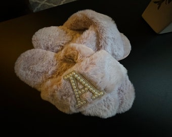 Può includere: Un paio di pantofole morbide di colore rosa chiaro con una grande lettera "A" incrostata di perle sulla parte superiore di una pantofola.