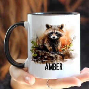 May include: White ceramic mug with a black rim and handle. The mug features a watercolor illustration of a raccoon standing on a rock in a forest setting. The text "AMBER" is printed in black at the bottom of the mug.