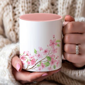 Op de afbeelding: Een witte keramische mok met een roze binnenkant en een bloemenontwerp met roze kersenbloesems op een tak. De mok wordt vastgehouden door een persoon die een witte trui draagt.