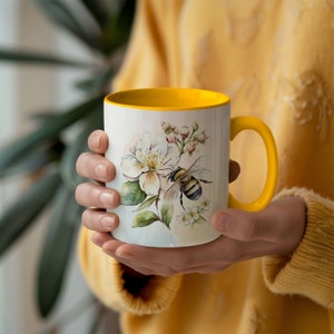 May include: White ceramic mug with a yellow interior and handle. The mug features a watercolor design of a bee, white flowers, and green leaves. The mug is held by a person wearing a yellow sweater.