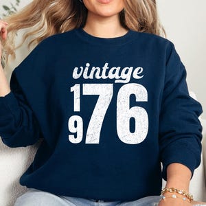 May include: Navy blue sweatshirt with the word "vintage" above the numbers "176" in white. The sweatshirt is a crew neck style. The image shows a person wearing the sweatshirt.