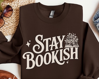 May include: A dark brown sweatshirt with the words "STAY BOOKISH" in a cream-colored, whimsical font. The design includes an open book with floral elements. The sweatshirt is a cozy, casual garment.