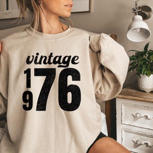 May include: A beige sweatshirt with the text "vintage 1976" in black. The word "vintage" is in a script font above the numbers. The numbers "1976" are in a bold, distressed font.