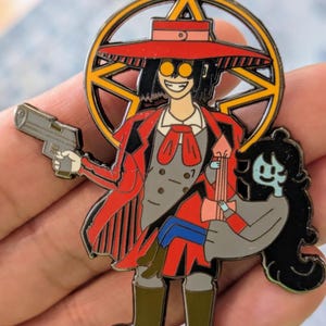 May include: A metal enamel pin featuring a character in a red coat, hat, and yellow glasses holding a gray handgun, standing in front of a yellow pentagram. The character carries a smaller blue-skinned figure with long black hair and a gray outfit.