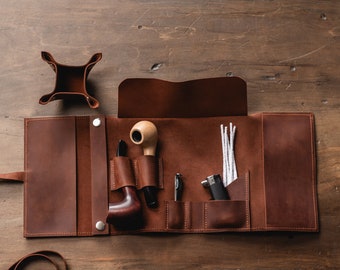 May include: A brown leather tobacco pipe roll-up kit with a pipe, tools, and a small tray. The kit is open, revealing a pipe, a tamper, a pen, a lighter, and pipe cleaners. The leather has a natural grain and is secured with snaps.