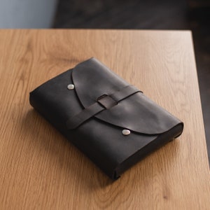 May include: A black leather journal with a flap closure and a silver metal buckle. The journal is closed and lying on a wooden surface.