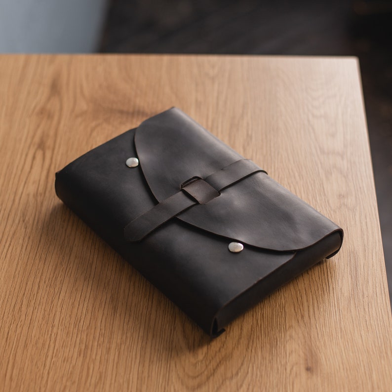 May include: A black leather journal with a flap closure and a silver metal buckle. The journal is closed and lying on a wooden surface.