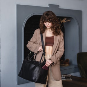 K&ouml;nnte beinhalten: Eine Frau in einem beige Blazer und einem braunen Oberteil h&auml;lt eine schwarze Ledertasche mit einem braunen Lederportemonnaie in der vorderen Tasche.