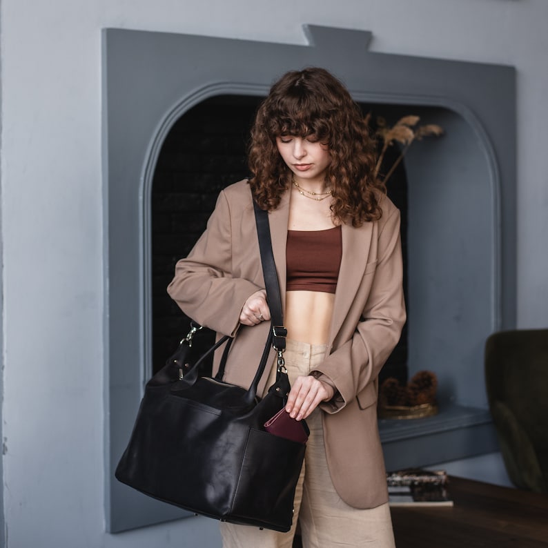 K&ouml;nnte beinhalten: Eine Frau in einem beige Blazer und einem braunen Oberteil h&auml;lt eine schwarze Ledertasche mit einem braunen Lederportemonnaie in der vorderen Tasche.
