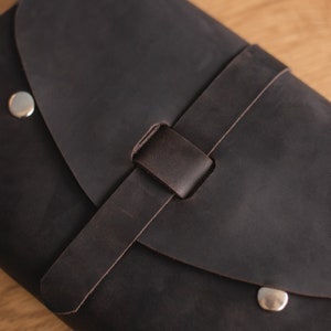 May include: A dark brown leather bag with a flap closure and a silver metal buckle. The bag has a strap that is folded over and secured with a silver metal button.