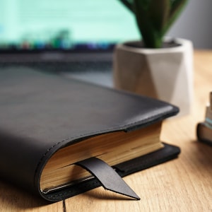 May include: A black leather-bound journal with a gold-toned page edge and a black ribbon bookmark. The journal is closed and lying on a wooden surface.