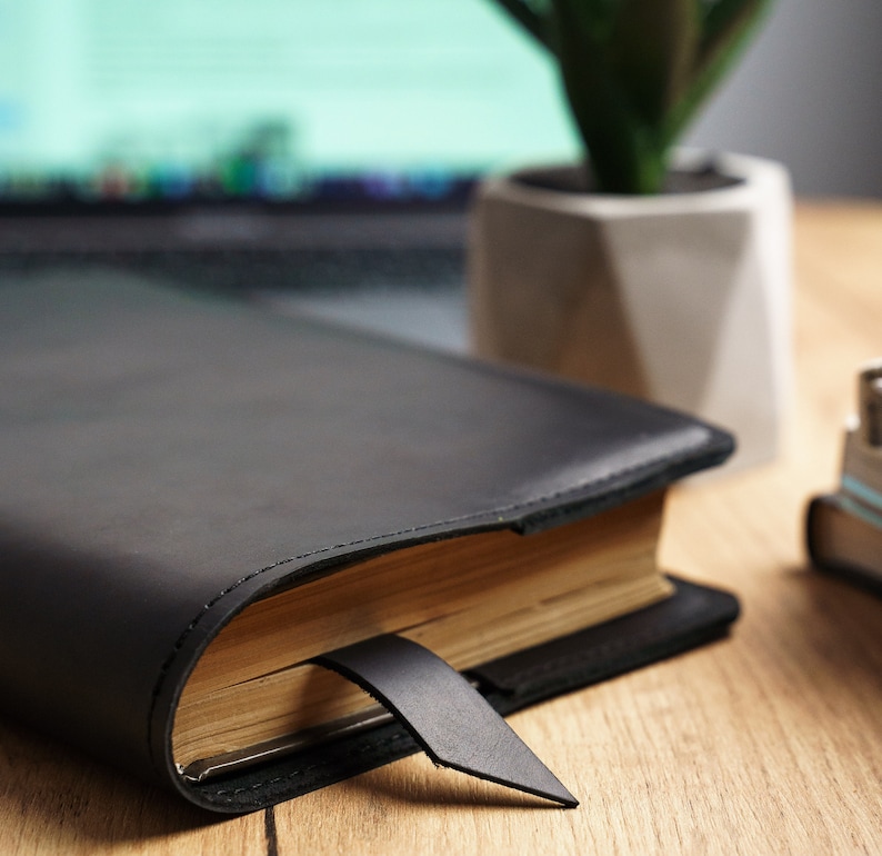 May include: A black leather-bound journal with a gold-toned page edge and a black ribbon bookmark. The journal is closed and lying on a wooden surface.