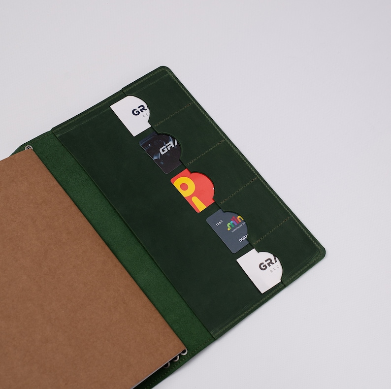 May include: A dark green leather notebook cover with card slots. The cover is open, revealing a brown paper notebook and several credit cards. The cards are visible in the slots, with various logos and text. The cover is on a white background.