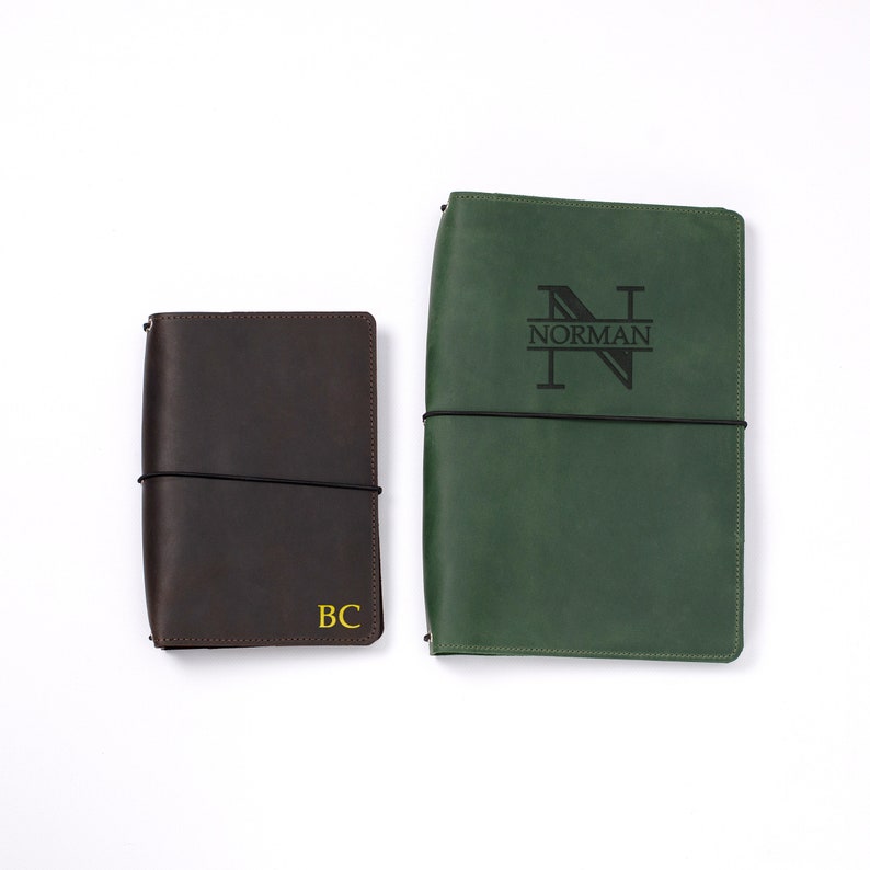 May include: Two leather journals, one dark brown and one green, lie side by side. The brown journal has the initials "BC" in yellow. The green journal is embossed with the name "NORMAN" and a stylized "N". Both have black elastic closures.