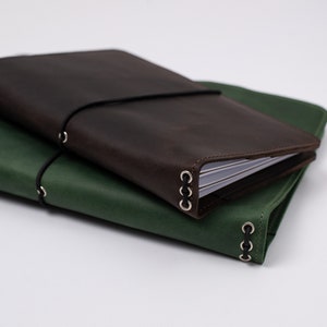 May include: Two leather journals, one dark brown and one green, are stacked on top of each other. Each journal has a black elastic band closure and metal ring accents. The journals appear to be new and unused.