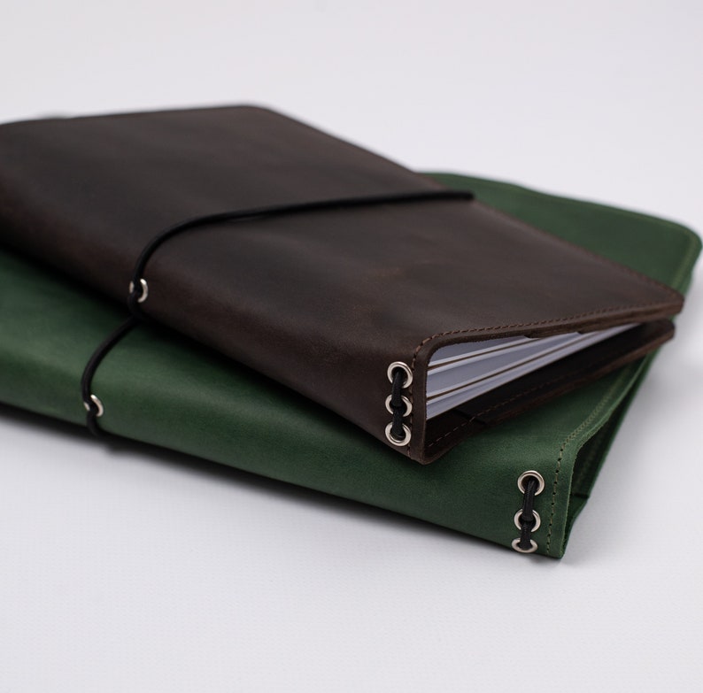 May include: Two leather journals, one dark brown and one green, are stacked on top of each other. Each journal has a black elastic band closure and metal ring accents. The journals appear to be new and unused.