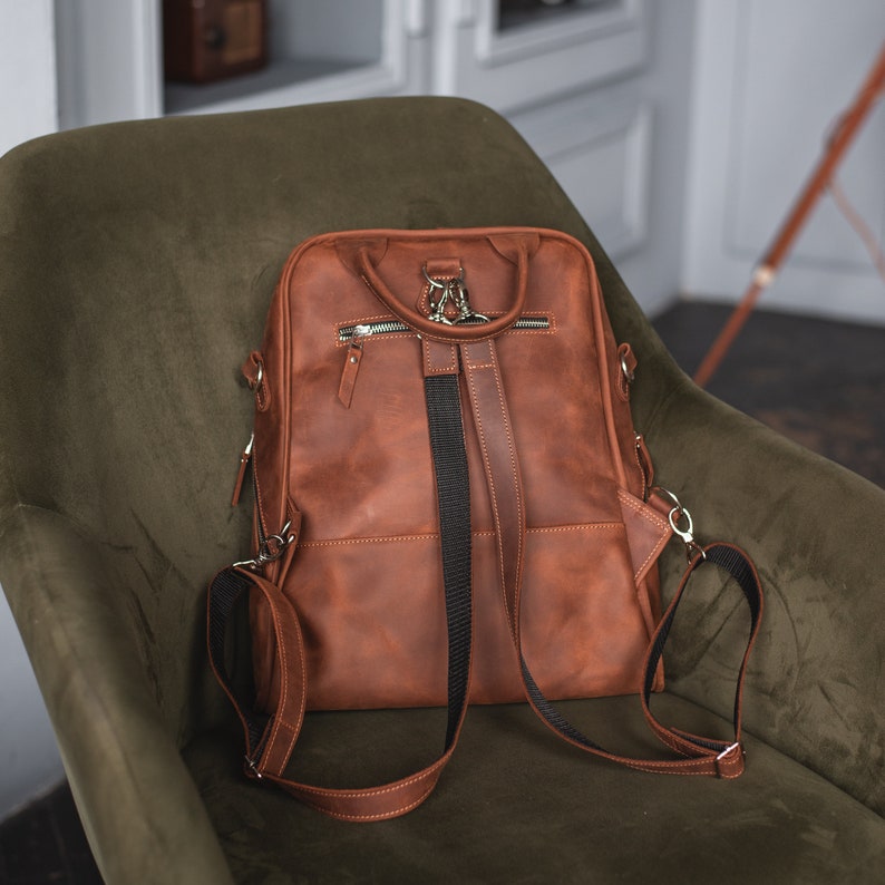 May include: A brown leather backpack with adjustable straps and multiple pockets. The backpack has a top handle and a zipper closure.