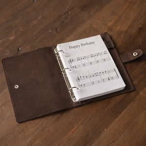 May include: Open brown leather binder with a sheet music insert. The sheet music is labelled "Happy Birthday" and includes musical notes for piano. The binder is open on a wooden surface.